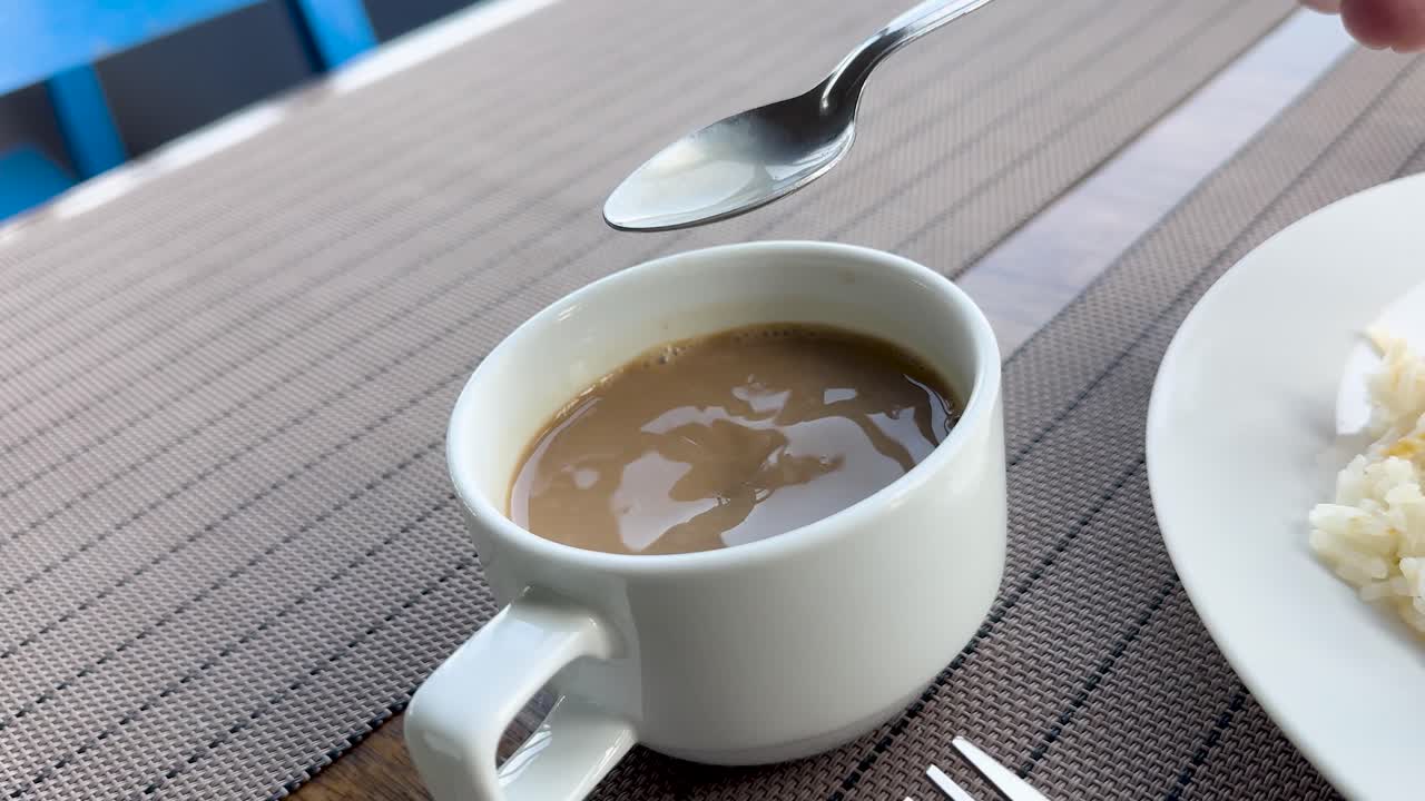 Hand stirs coffee in white cup on patio table, natural daylight, steady medium close-up shot