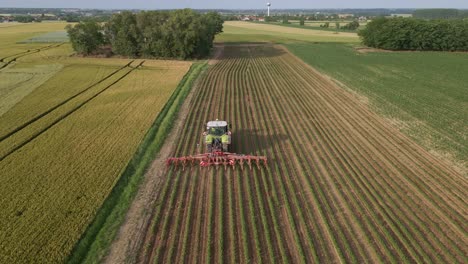 a tractor is seen from above as it moves along neatly arranged rows of crops on a cultivated field. the surrounding landscape features patchwork farmland, trees, and a distant village.