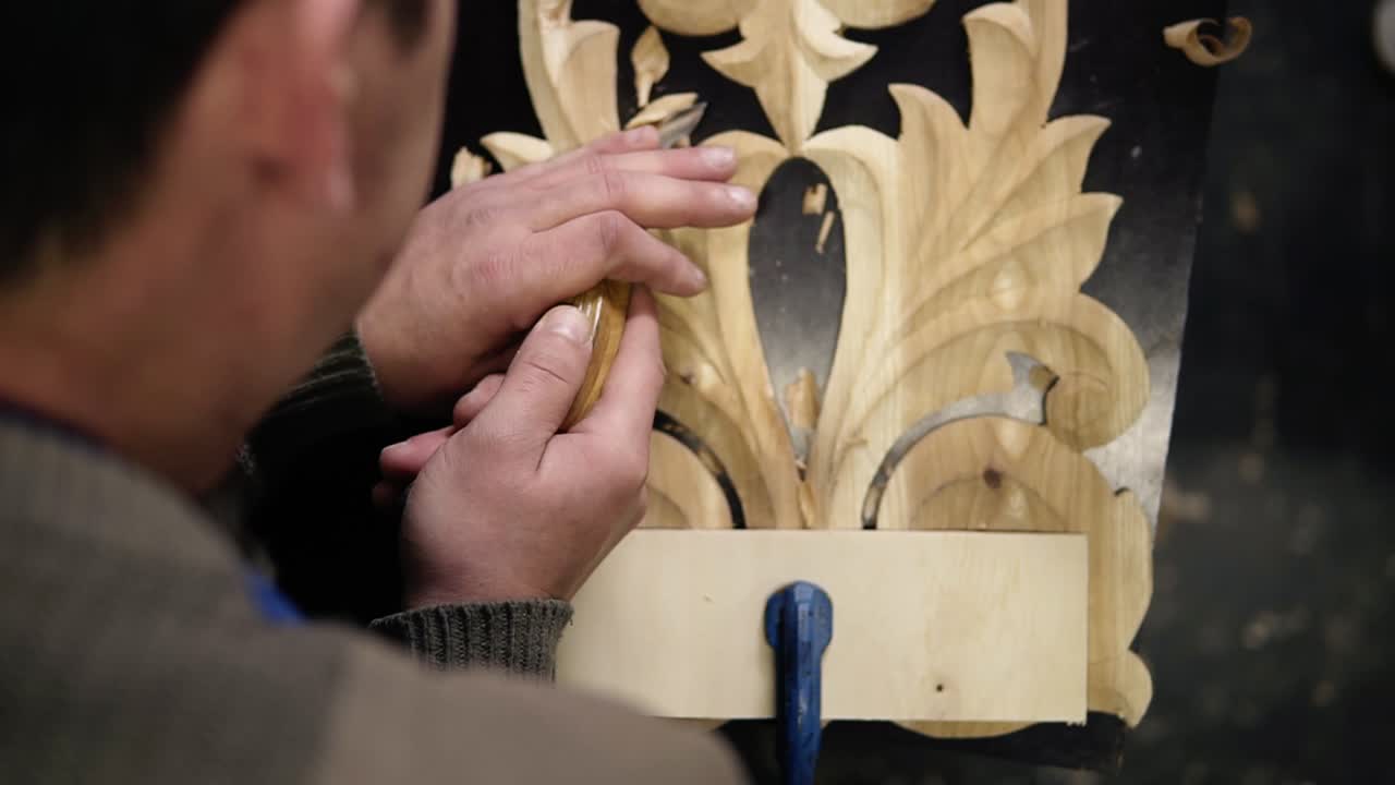 Close up footage of man's hands, he is curving a small details on the piece wodden pattern - floral ornament in work shop on the work table using small planer. Footage from the top