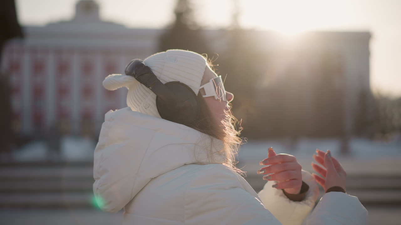 Close up of student wearing winter coat, dancing with visible breath in cold air while sunlight flares in background, headphones on, relaxed expression, joyful winter vibe captured in sunset