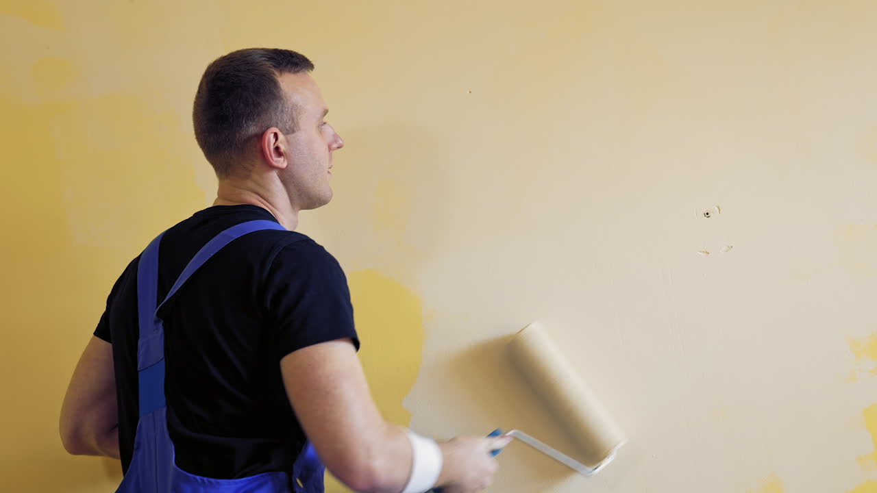 Male worker with a paint roller. Young man in worker's suit painting wall in light color. Professional painting services.