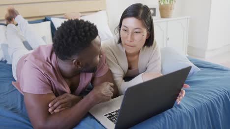 Happy diverse couple using laptop and lying in bedroom
