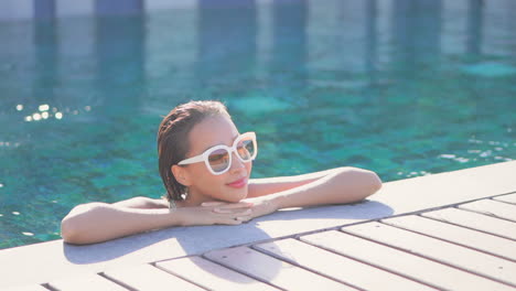 Retrato-De-Una-Hermosa-Mujer-Asiática-Con-Gafas-De-Sol-Blancas-Sumergidas-En-Una-Piscina-De-Agua-Con-La-Barbilla-En-Las-Manos-Cruzadas