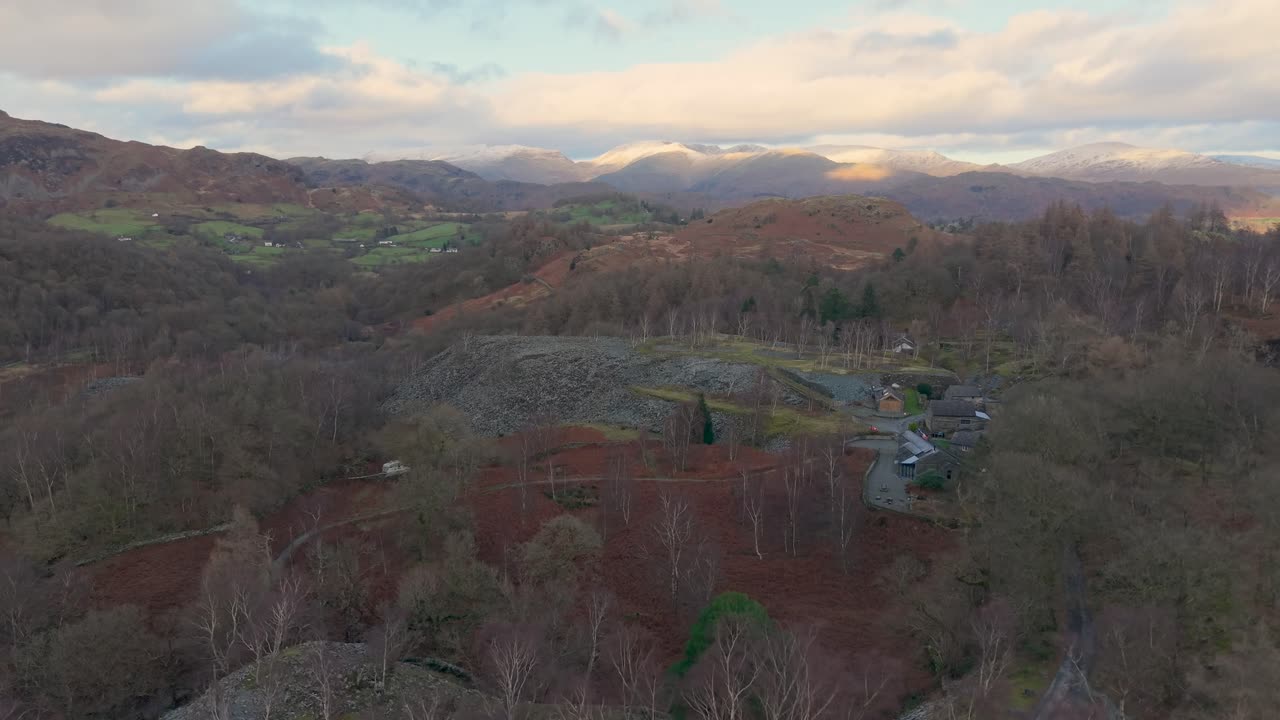 A drone ascends over slate ground, revealing distant hills and a mountain glowing under sunlight breaking through clouds, expanding the scenic Lake District landscape ahead