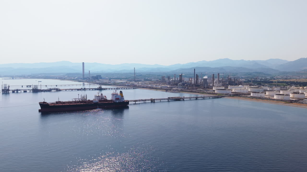 Drone panning sideways over a red oil tanker docked at a pier near a refinery, with sea and mountains in the background