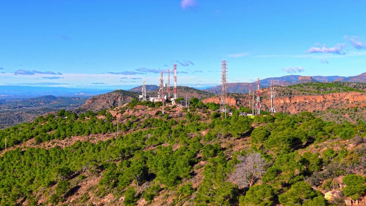 Gruppo di torri di telecomunicazione su un crinale montuoso boscoso, regione di Valencia, Spagna. Palo dell'antenna rosso e bianco che si erge sopra una pineta e una scogliera rocciosa di arenaria sotto un cielo azzurro.
