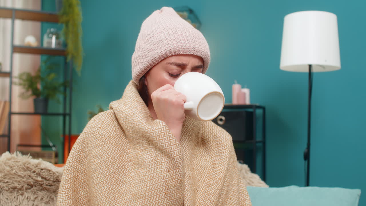 Giovane donna sotto una coperta sul divano, con un cappello di lana, che beve tè caldo combattendo un brutto raffreddore con pazienza.