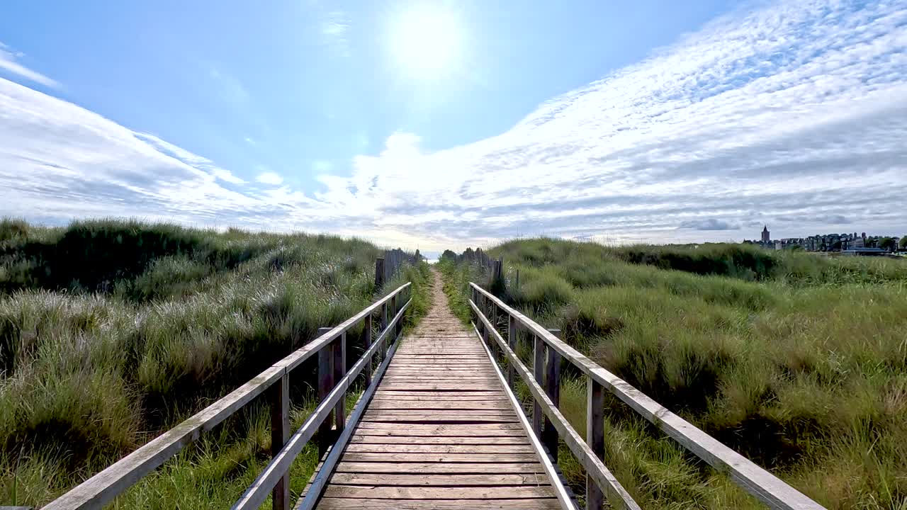 Forward motion along sunlit wooden boardwalk, grassy dunes, blue sky, and clouds in Fife, Scotland