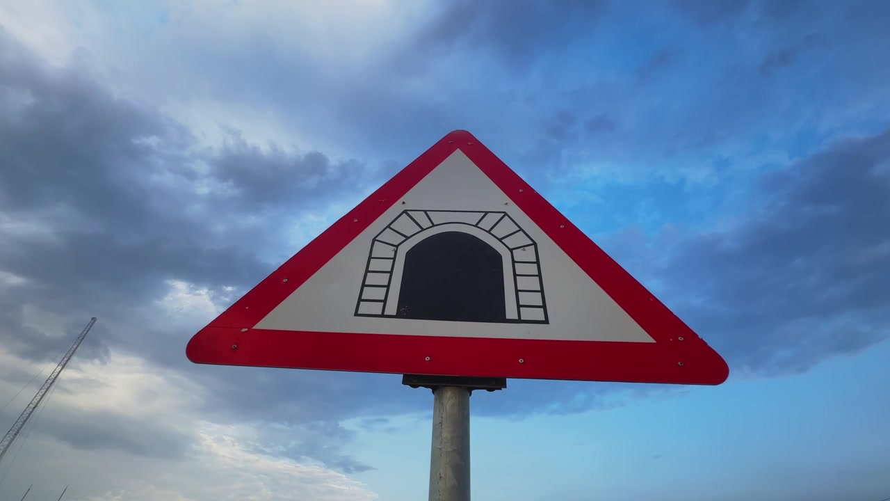 Dramatik bulutlu bir gökyüzüne karşı üçgen bir tünel uyarı yol levhasının etrafında dönen kamera, ulaşım güvenliğini, yol kenarı tabelalarını ve otoyol navigasyon altyapısını vurguluyor.