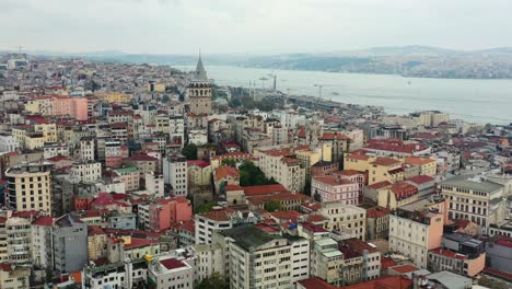 aerial drone revealing galata tower in surrounded by old european residential buildings on a cloudy day in istanbul turkey with the bosphorus river in the distance