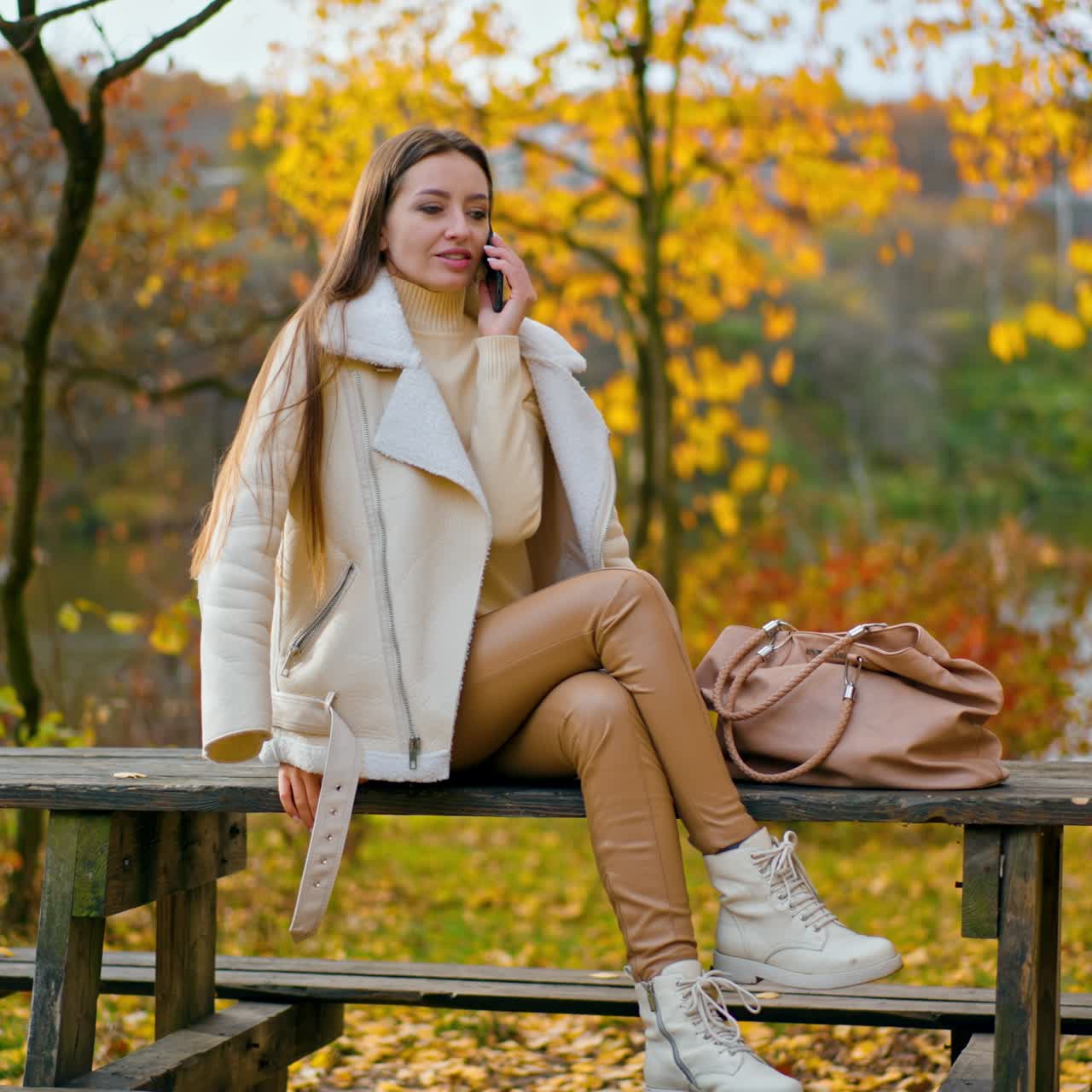 Attraktive, langhaarige Brünette telefoniert im Freien. Dame in warmer Kleidung sitzt auf dem Tisch in der wunderschönen herbstlichen Natur