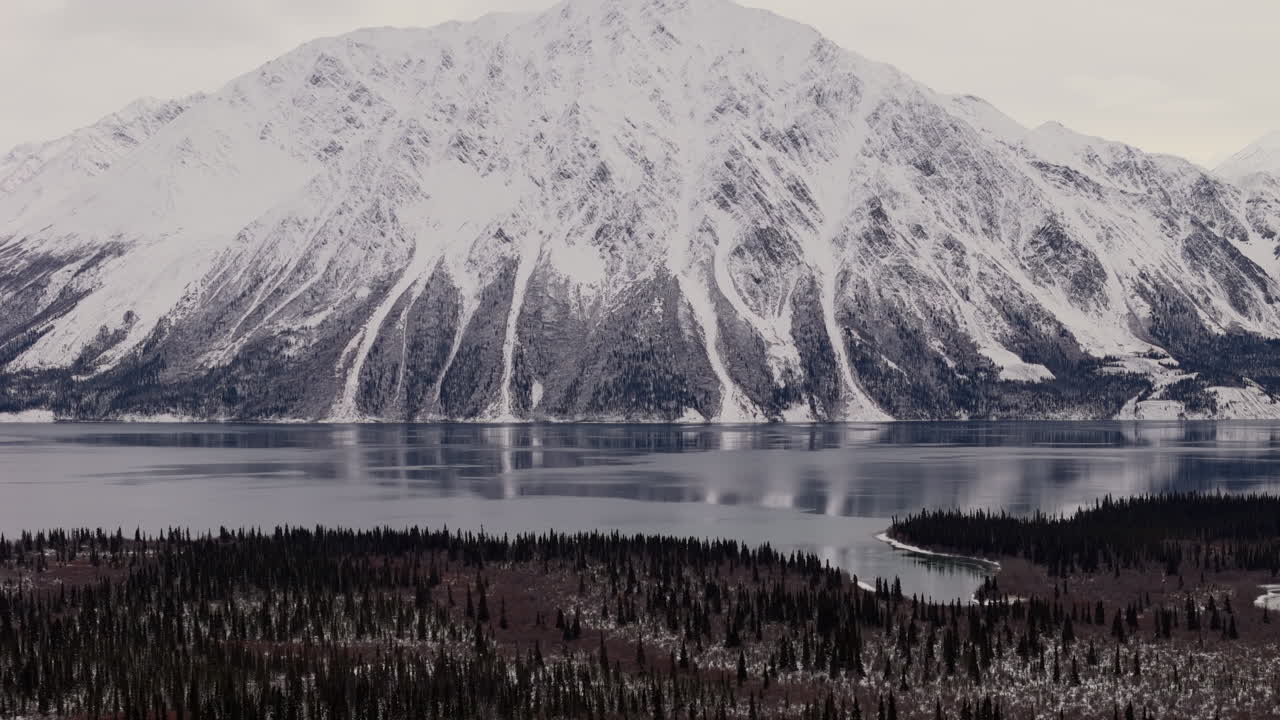 Panorama di montagne innevate, foresta e il Lago Kathleen ghiacciato nello Yukon, Canada. - Ripresa aerea con tilt-up.