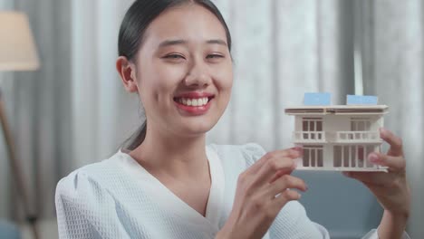 웃는 아시아 여성 엔지니어가 태양광 패널을 가진 작은 집 모델을 들고 집에서 데스크에서 일하는 동안 카메라에 엄지손가락을 보여주고 있습니다.