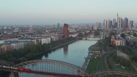 Empty-Cargo-Train-going-over-Bridge-on-Main-River-in-Frankfurt-am-Main-with-Skyscraper-Skyline-view,-Early-morning,-Aerial-sideways-left