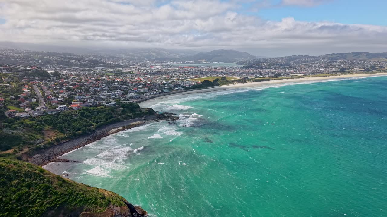 Drone shot moving forward along St Clair Beach showing turquoise waves and the coastal city of Dunedin in the background