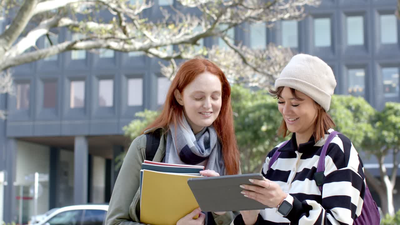 Kvinnliga vänner lutar sig vid en gångväg på campus, upptäcker nytt innehåll på en surfplatta, delar anteckningar och skrattar.