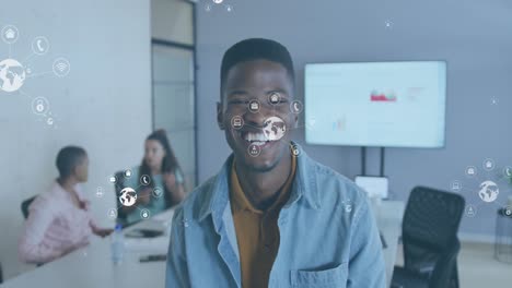 Animation-of-multiple-icons-around-globes-over-smiling-african-american-man-standing-in-office