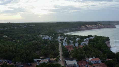 Tilt-up-aerial-drone-shot-of-the-main-street-leading-into-the-famous-tropical-tourist-town-of-Pipa,-Brazil-in-Rio-Grande-do-Norte-with-large-cliffs-above-the-calm-turquoise-ocean-and-green-foliage