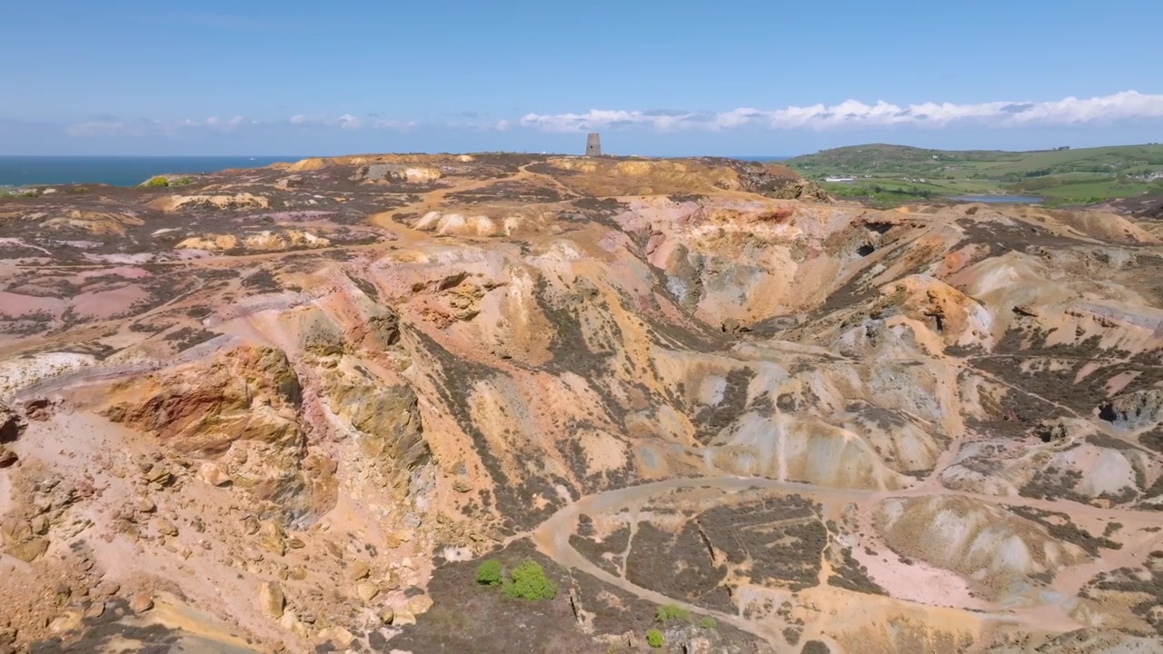 Miniera di rame di Parys Mountain, Amlwych, Anglesey, Galles, Regno Unito. Volo di drone inverso che si allontana da un mulino a vento abbandonato in cima a una collina, rivelando un profondo cratere e colori minerali.