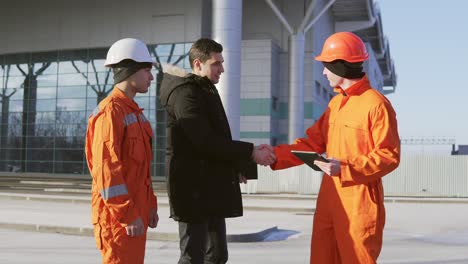 investor of the project in a black suit examining the building object with construction workers in orange uniform and helmets.