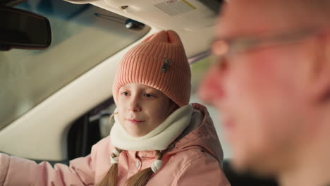 young girl, dressed warmly in a pink beanie and jacket, is sitting in the front passenger seat of a car, engaging in conversation or activity with a man seated beside her. the man, possibly her father