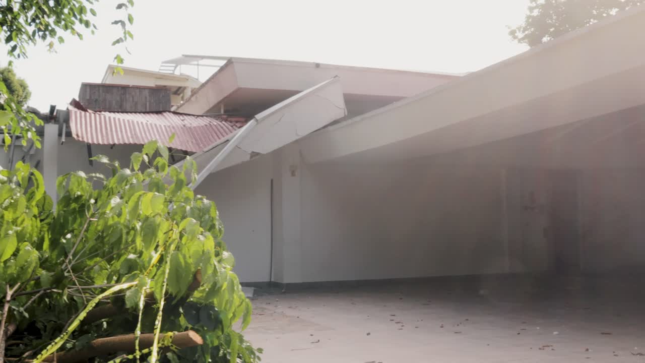 Side façade of an abandoned bungalow home with peeling paint, falling asphalt roof, fallen tree and eerie front porch stands out on a quiet rural highway.