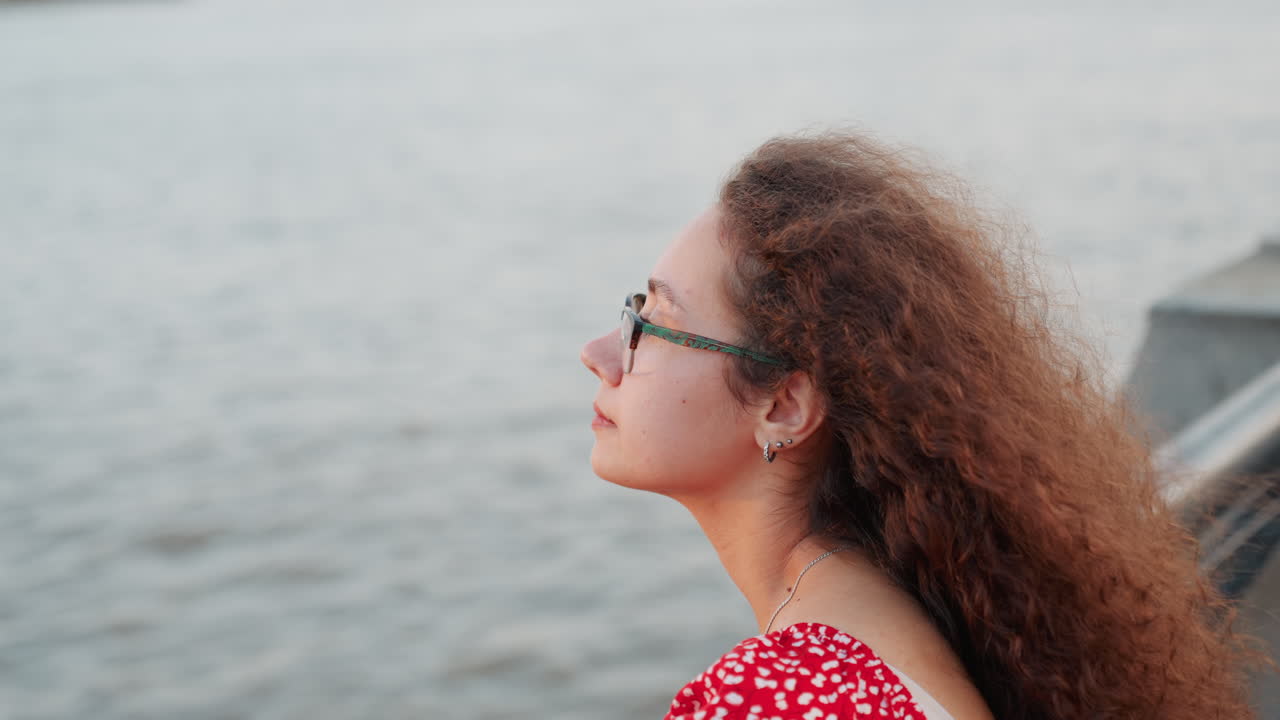 Young woman gazing at water, student on riverside railing with long curly hair and glasses in red polka dress, soft evening light, calm river background, introspective mood and gentle breeze