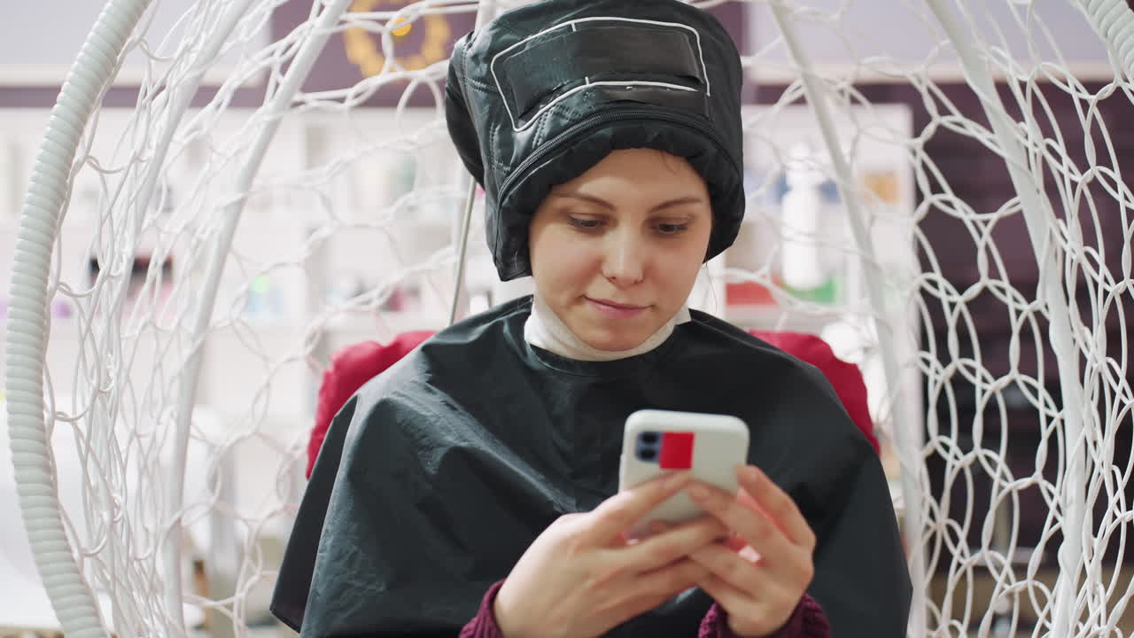 Kafkaslı bir kadın, kuaförde başlığı takılı kurutucunun altında, pelerin içinde rahat bir pozisyonda, telefon görüşmesi sırasında salıncakta otururken telefona gülümsüyor, yumuşak stüdyo ışığı, arka planda kuaför hareketliliği, güzellik seansı sırasında keyifli bir chat ve iletişim