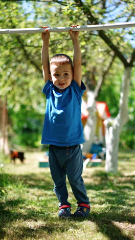 three-year-old boy doing exercises on the horizontal bar. cute kid hanging on the bar lifting his feet. sunny summer day in the garden at backdrop. vertical video.