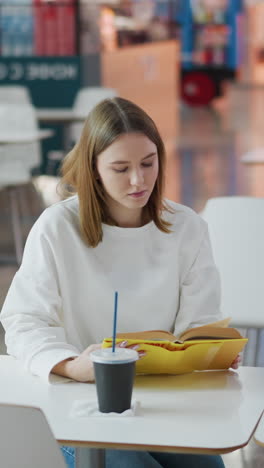 joven sentada sola en una mesa en el centro comercial, leyendo su libro con enfoque tranquilo, fondo suave cuenta con asientos vacíos, una bebida y bolsas de compras en la mesa