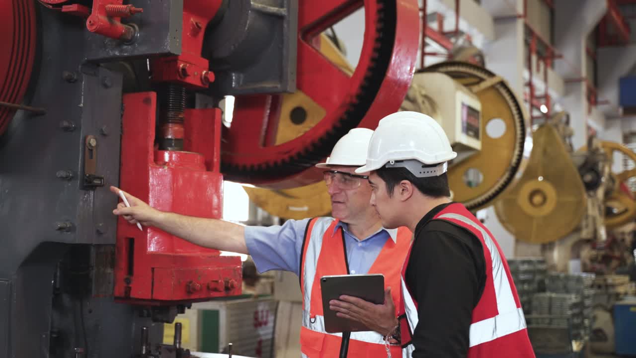 Senior man engineer wearing safety goggles and hard hat standing holding tablet with training young engineer operate hydraulic press stamp machine in factory.