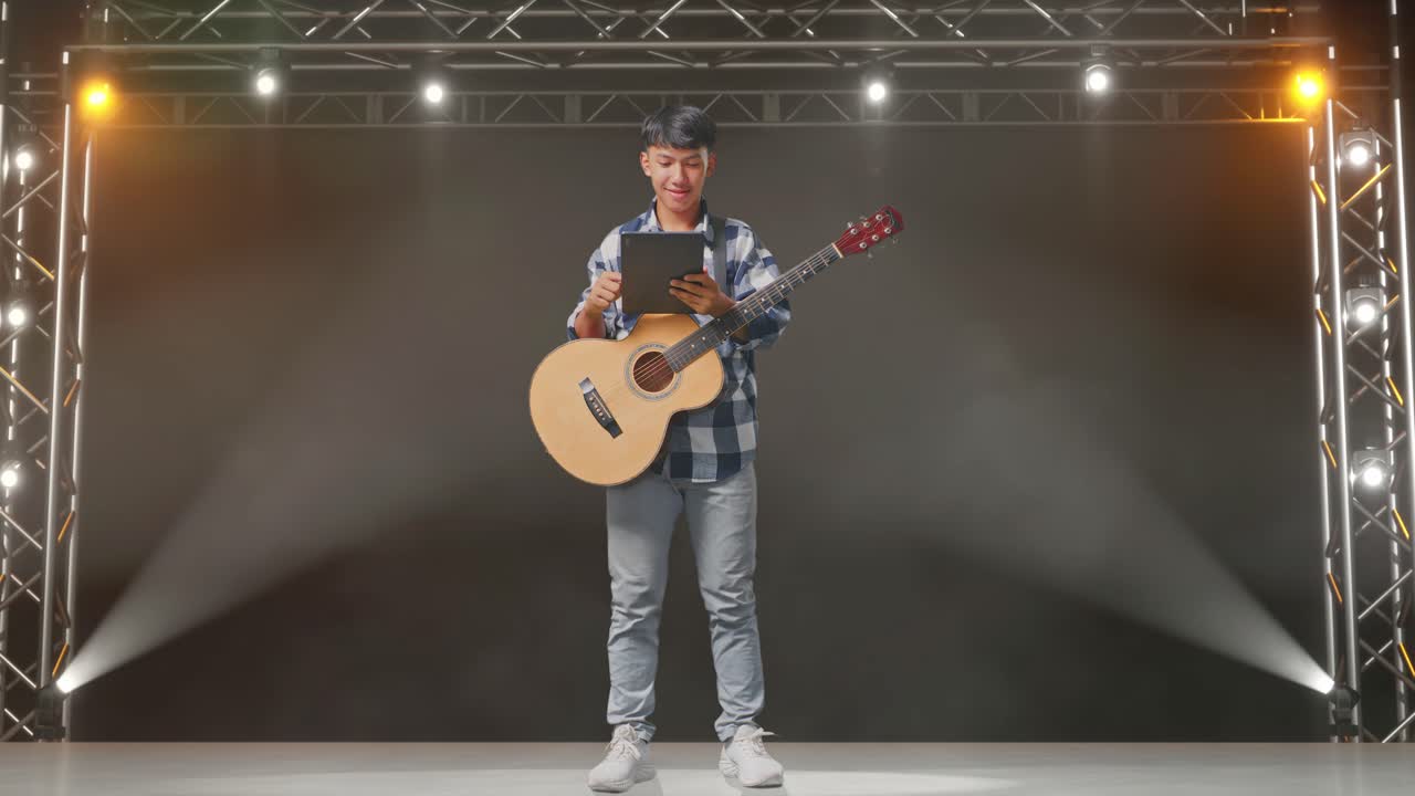 cuerpo lleno de adolescente asiático con guitarra usando una tableta mientras está de pie en el escenario