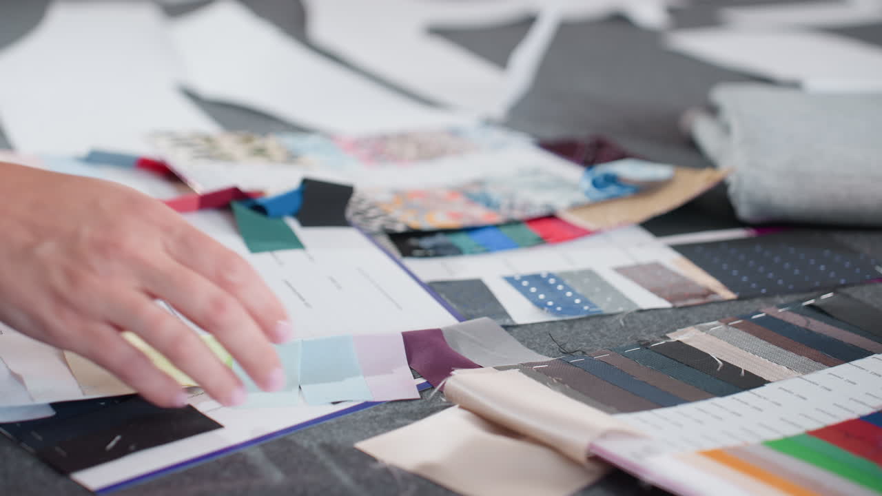 Close up view of seamstress lifting colorful panel from fabric samples spread across work table, showing vibrant swatches, material options, design planning, and fabric organization