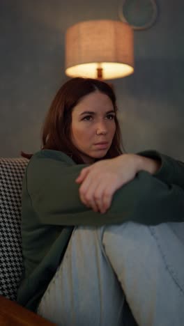 vertical video of a sad confident brunette girl in a green shirt pressing her legs to her body and sitting on a chair near a yellow lamp at home