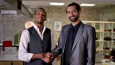 two multy-ethnic workers are shaking hands in office, watching at camera, smiling, communication concept