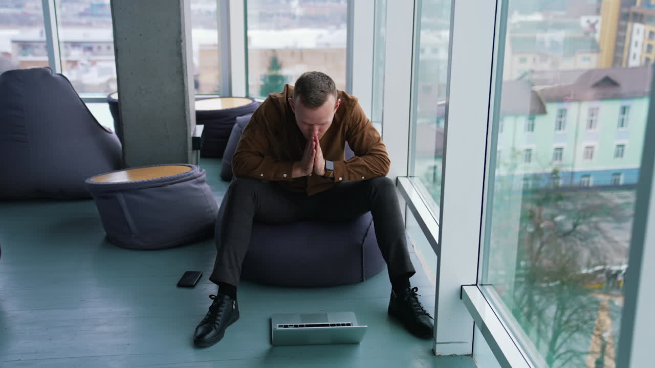 Young man with a laptop near the window. Handsome guy in shirt is sitting on a comfortable ottoman and looking on his laptop waiting for something.