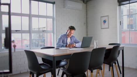 Focused-biracial-businessman-using-laptop-and-taking-notes-at-table-in-meeting-room,-in-slow-motion