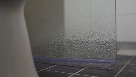 piso húmedo del baño pavimentado con azulejos grises detrás de la puerta de la ducha con gotitas que caen del cabezal de la ducha, vista panorámica lenta