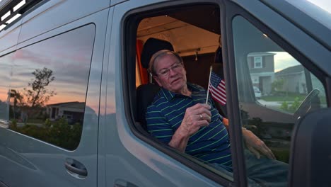 elderly man holds the american flag and waves it while sitting in his camper van