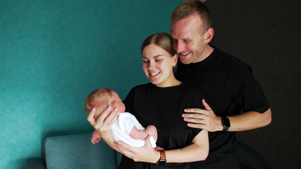 Woman holding a tiny newborn in hands kissing her husband on the cheek. Happy family of three having great time.