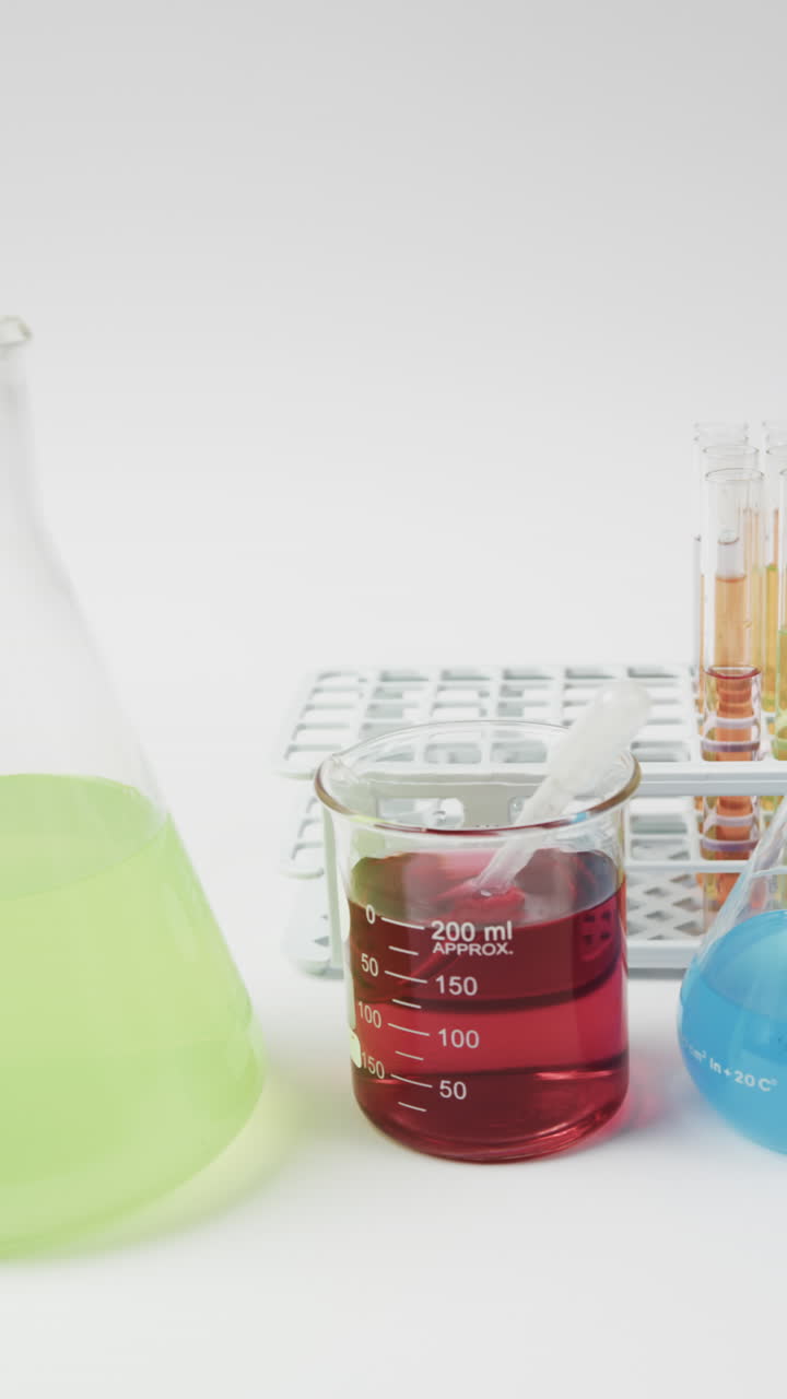 Vertical video of coloured liquids in flasks with test tubes in stand on white background