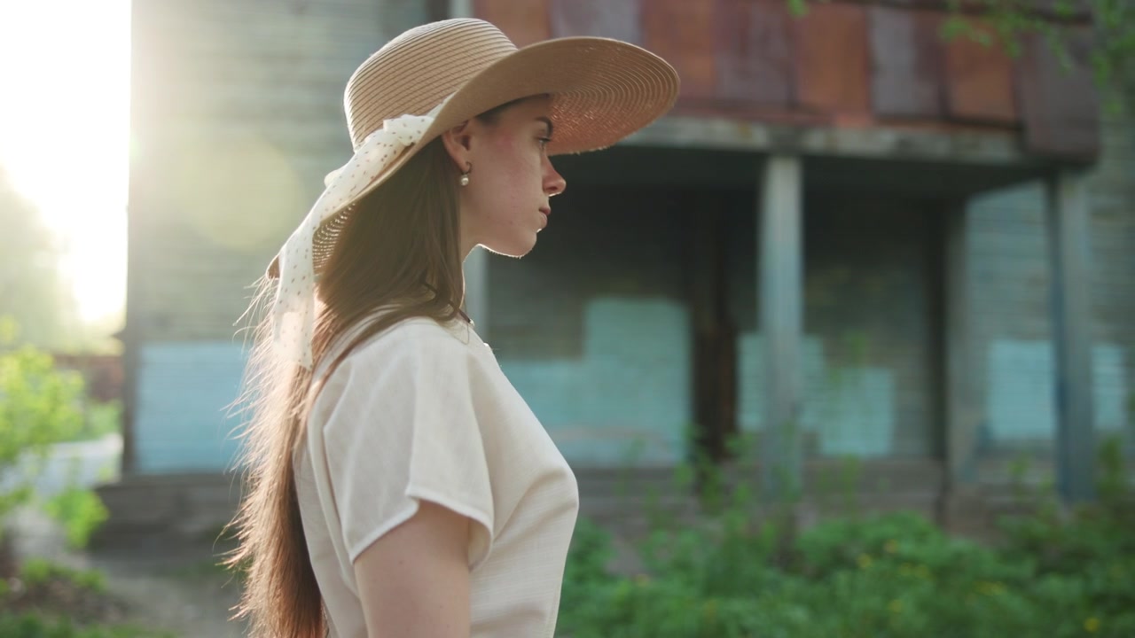 A young woman wearing a wide-brim hat is shown in side profile outdoors in warm sunlight, with a softly blurred building and greenery in the background