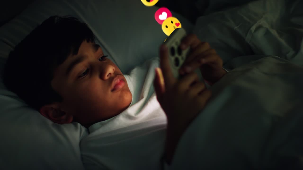 Young Boy Lying In Bed At Home At Night Looking At Mobile Phone With Motion Graphics Emojis Showing Multiple Social Media Or Messaging Notifications Liking And Reacting To Online Content