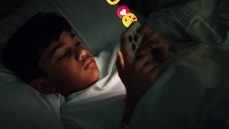 young boy lying in bed at home at night looking at mobile phone with motion graphics emojis showing multiple social media or messaging notifications liking and reacting to online content