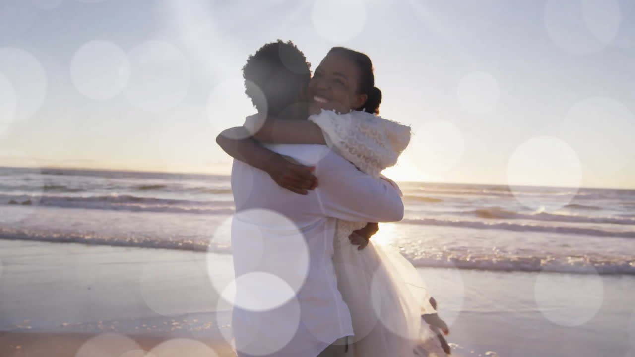 Animation of light spots over happy african american bride and groom embracing on beach at wedding
