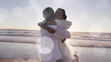 Animation-of-light-spots-over-happy-african-american-bride-and-groom-embracing-on-beach-at-wedding