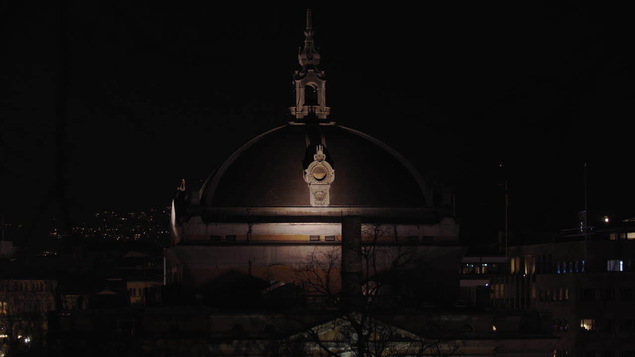 Cinematic slow motion close-up to wide 4K shot with parallax motion of the illuminated dome on top of the national theatre with tree branches moving in front of building, at night in Oslo Norway