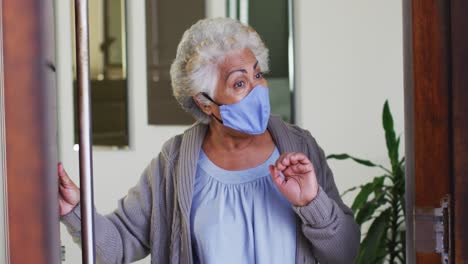 African-american-senior-woman-wearing-face-mask-opening-front-door-and-welcoming-at-home