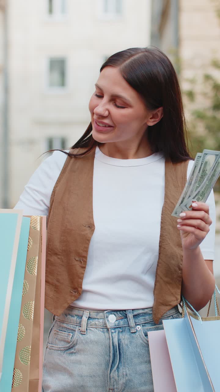 Happy young shopaholic caucasian woman holding cash while carrying shopping bags on city street