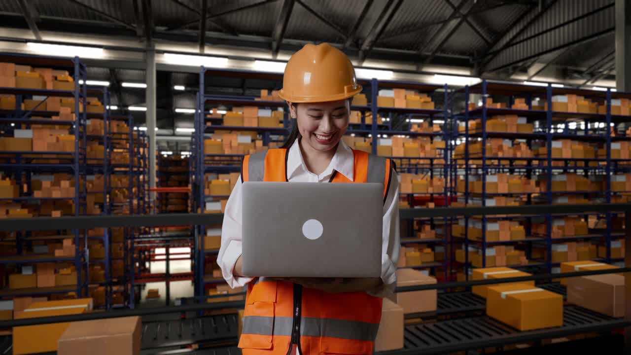 ingeniera asiática con casco de seguridad trabajando en una computadora portátil mientras está de pie en el almacén con estantes llenos de productos de entrega